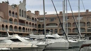 Un puerto deportivo con varios veleros amarrados, un edificio con balcones arqueados y un paseo pavimentado al frente.