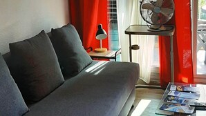 Een moderne woonkamer met een grijze bank, een bijzettafeltje met tijdschriften, een staande ventilator en een raam met rode gordijnen.