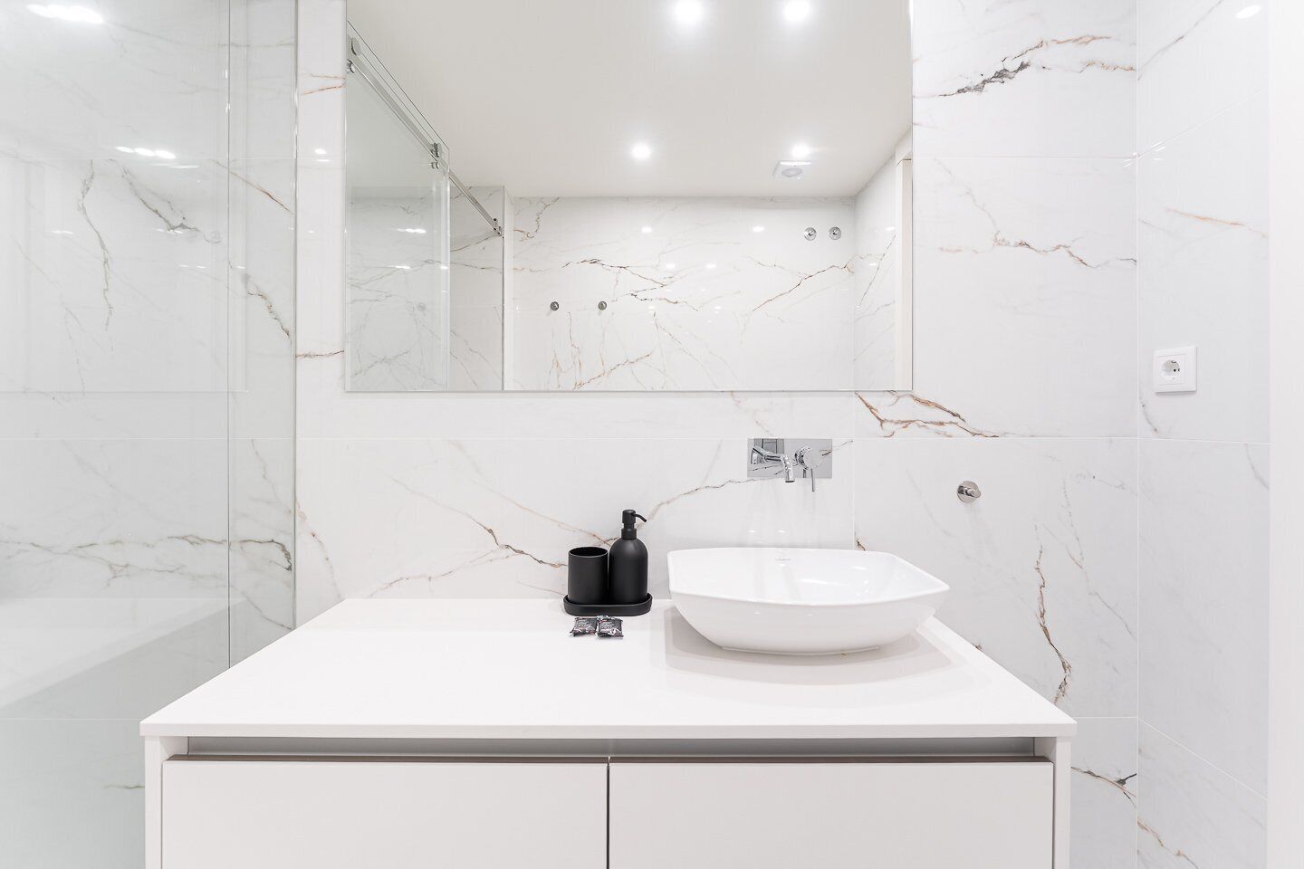 Un bagno moderno con un lavello in marmo bianco, un dispenser per sapone nero e un box doccia in vetro.