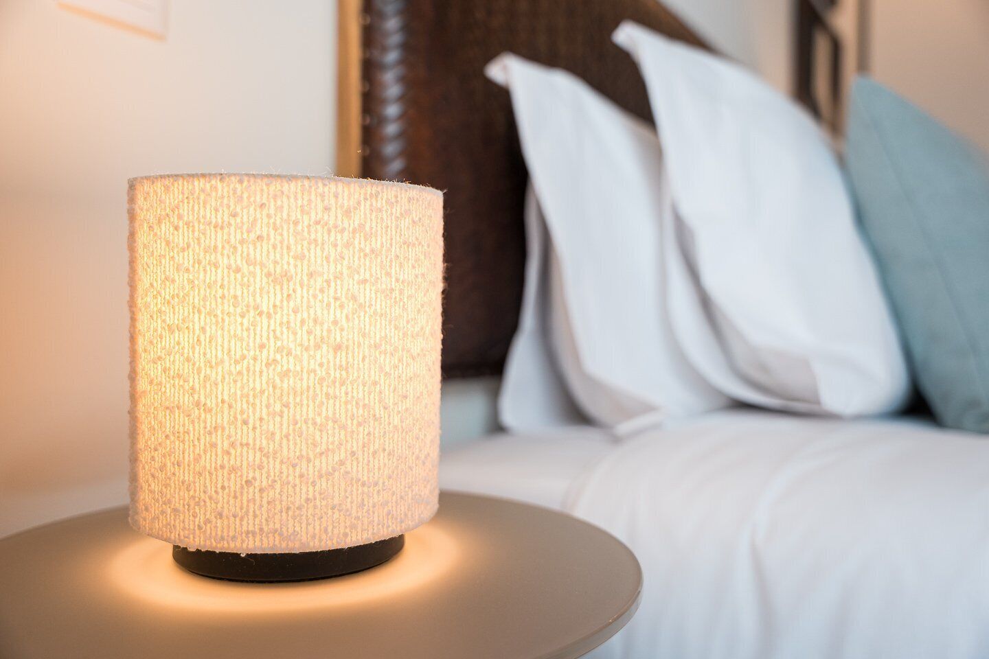 Une lampe cylindrique à la texture particulière, posée sur une table ronde dans une chambre d’hôtel, avec un lit et des oreillers en arrière-plan.