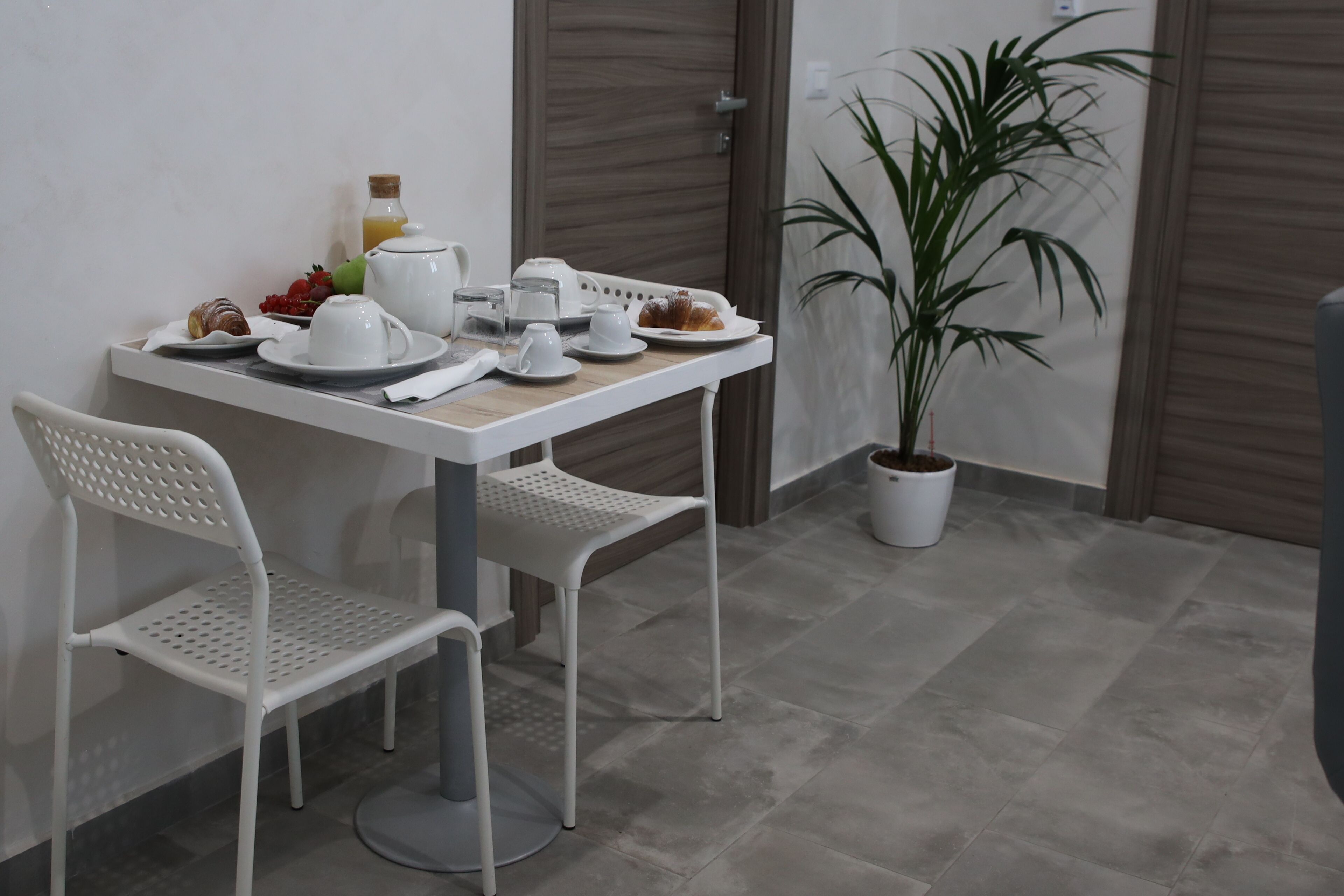 A dining area with a table set for two, including plates, cups, and a bottle of juice. There are two white chairs and a potted plant.