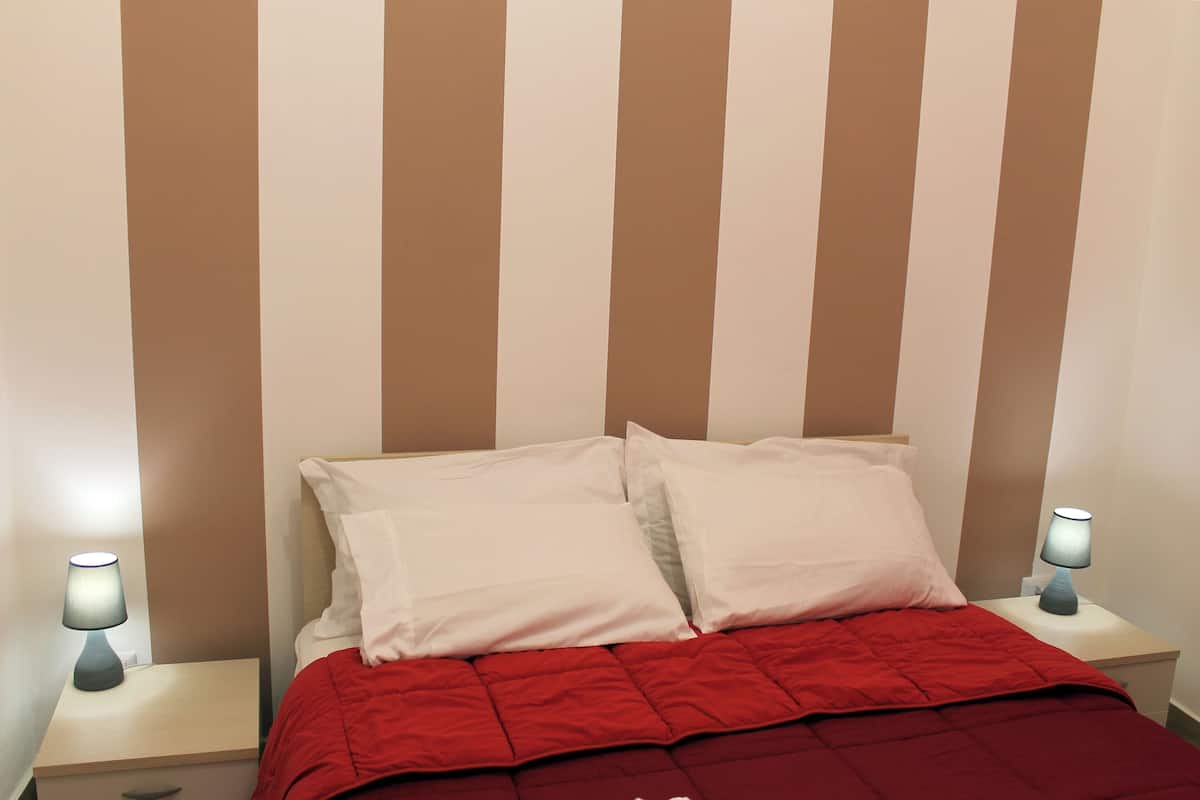 Una cama con almohadas blancas y una colcha roja, dos lámparas de noche y una pared a rayas.