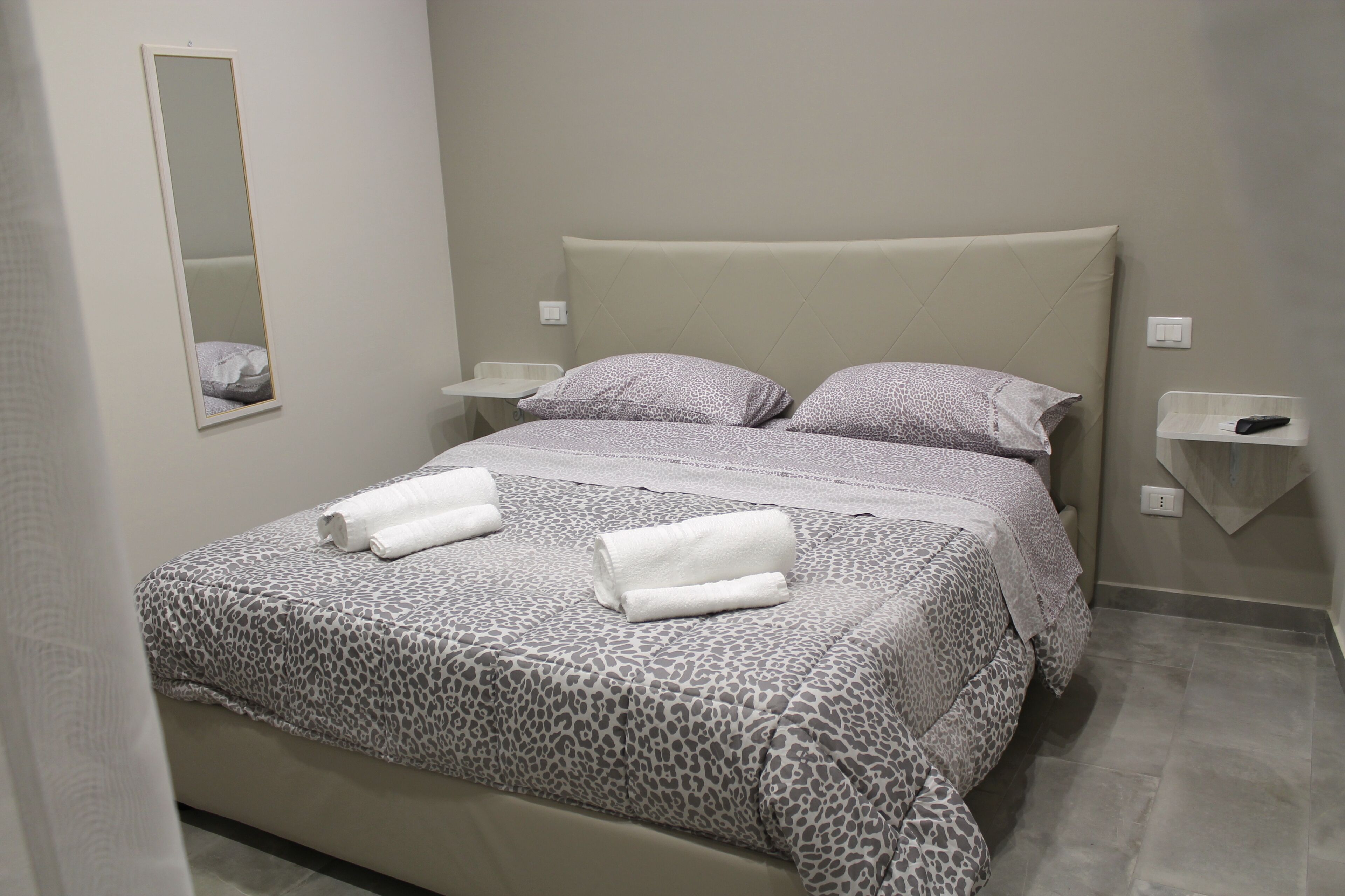 A neatly made bed with a leopard print bedspread and pillows, a bedside table with a remote control, and a wall-mounted mirror.