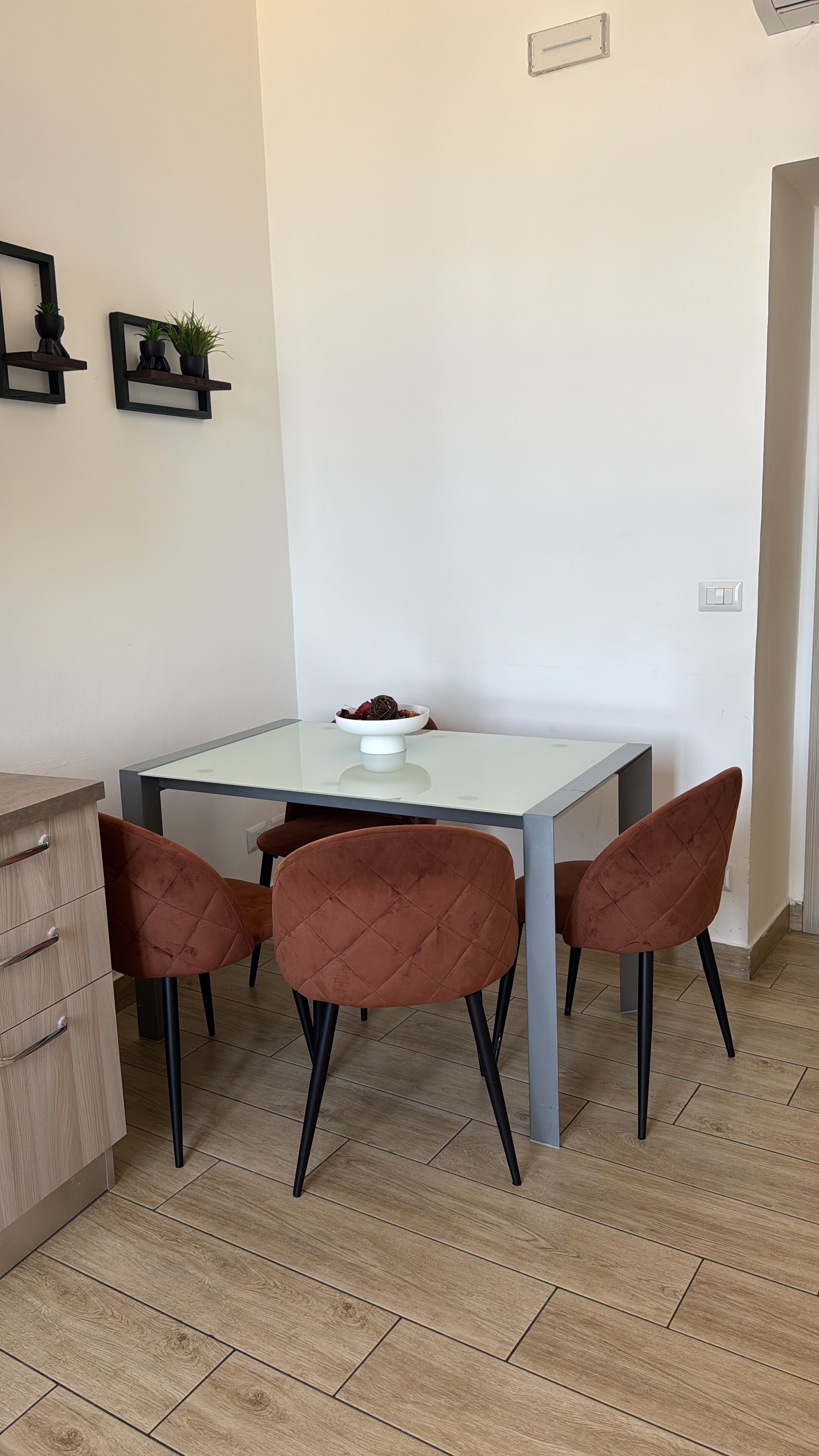 A small dining area with a glass-top table, two brown chairs, a wooden cabinet, and two wall-mounted shelves with plants.