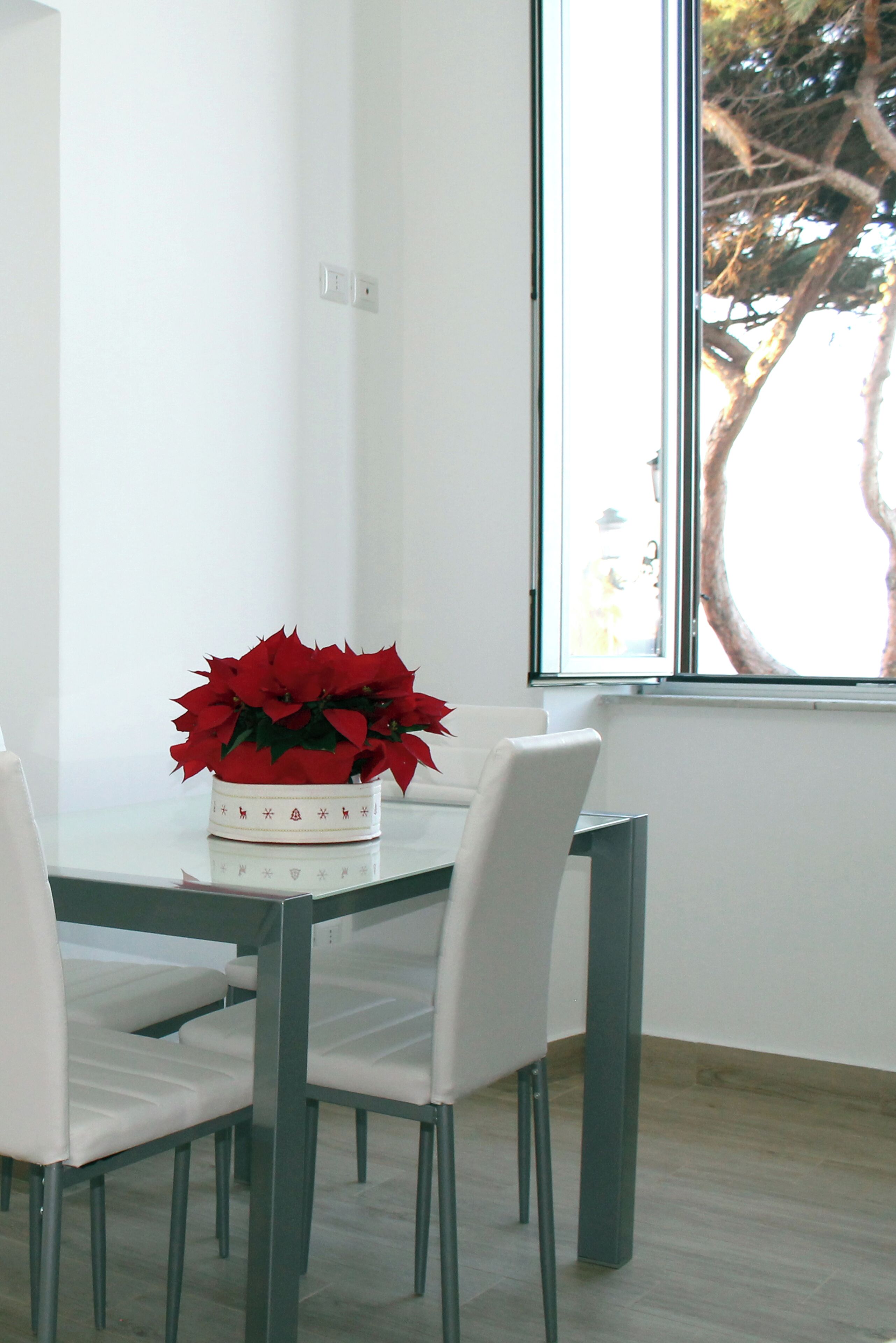 A dining area with a table and chairs, a vase with red flowers, and a window with a view of trees.