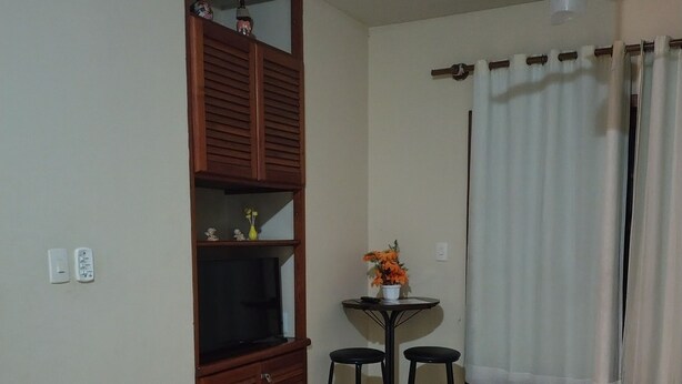 Una habitación con ventilador de techo, una mesita con dos taburetes, televisor sobre un mueble y una ventana con cortinas.