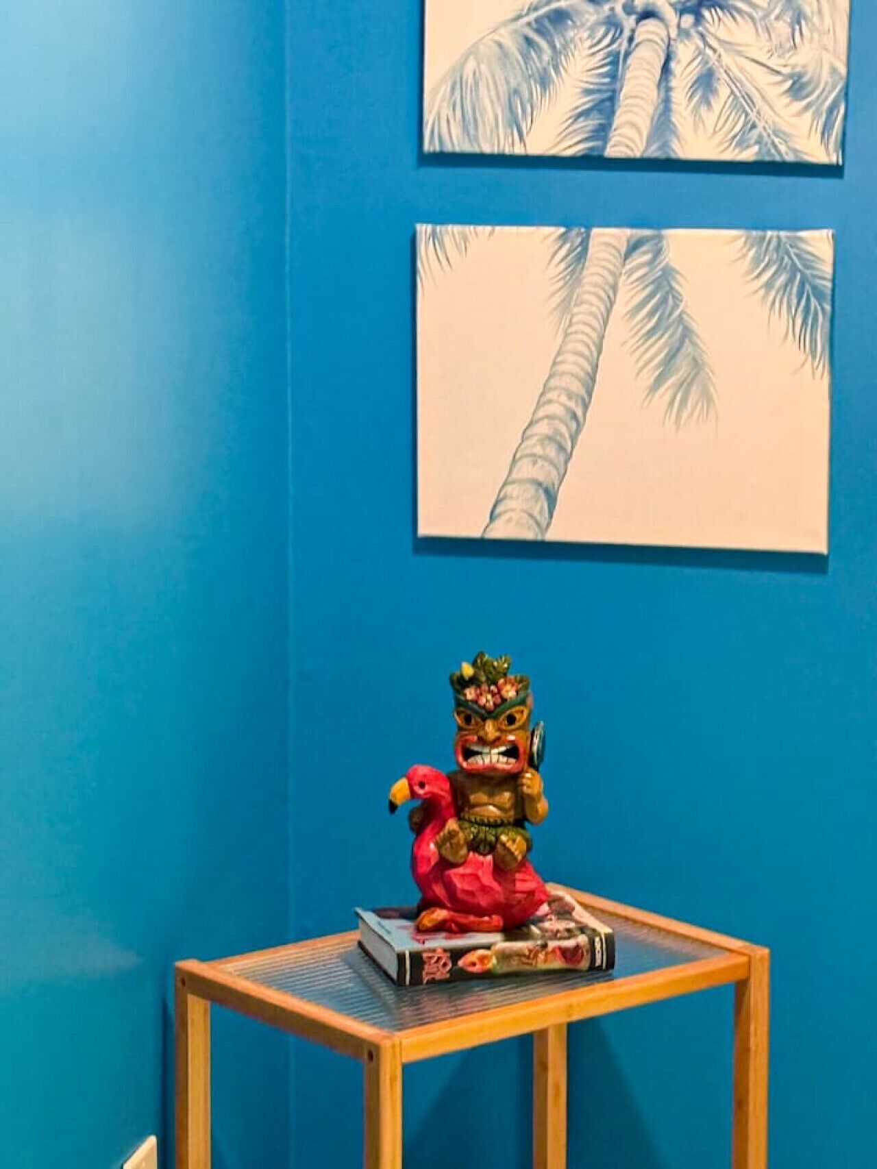 Una habitación con una pared azul, una pequeña mesa de madera con una estatua y un libro, y dos cuadros con imágenes de palmeras.
