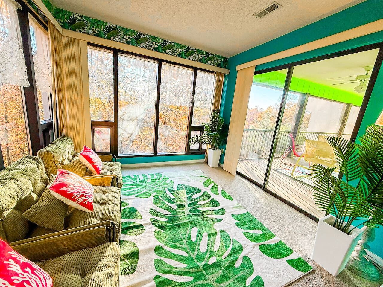 A sunlit room with a large window, a sofa with cushions, a tropical leaf-patterned rug, and a potted plant.