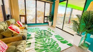 Une pièce avec une grande fenêtre, un canapé avec des coussins, un tapis à motifs de feuilles vertes et une plante en pot.