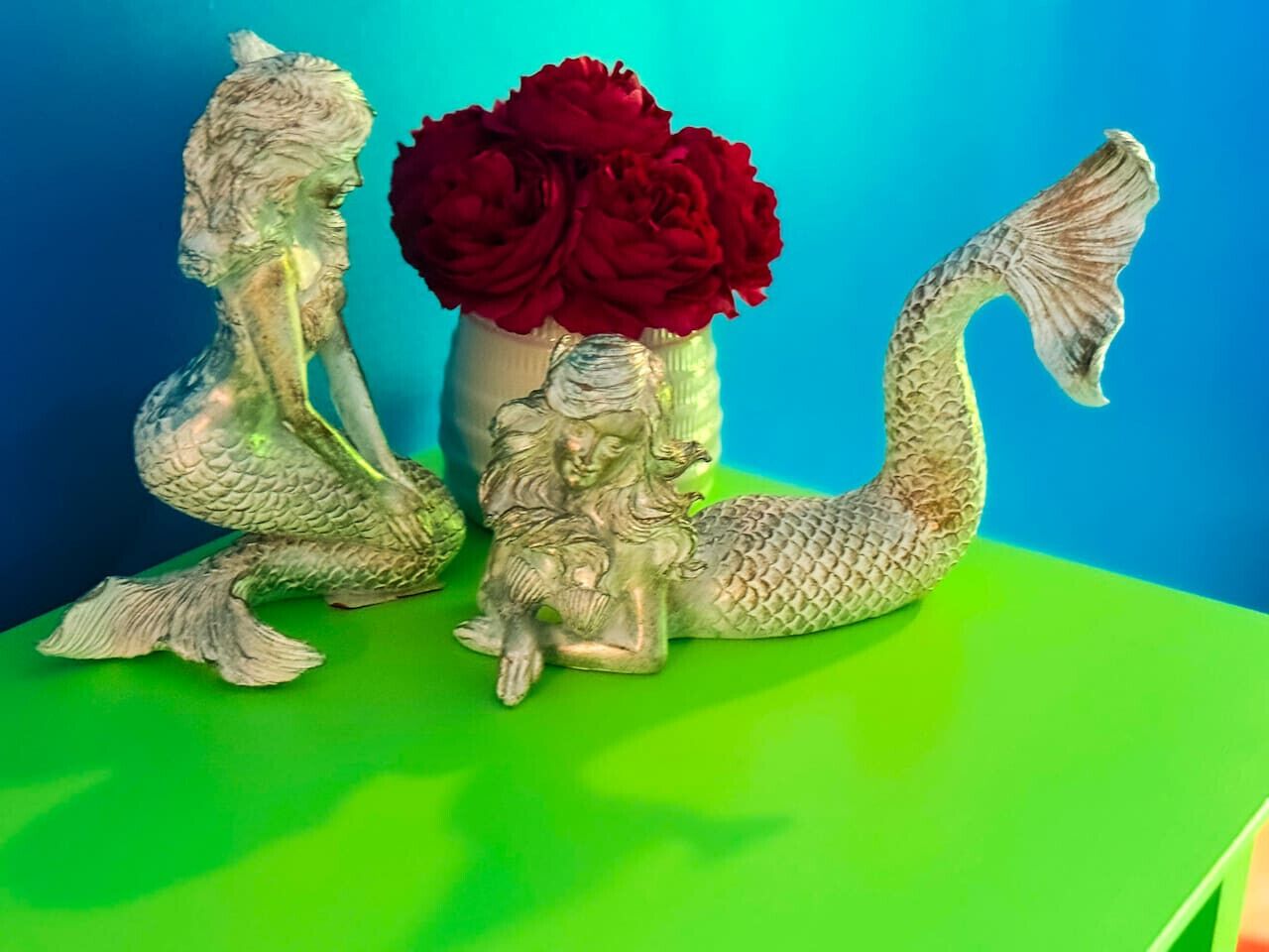 A mermaid figurine and a vase with red flowers on a green surface against a blue background.