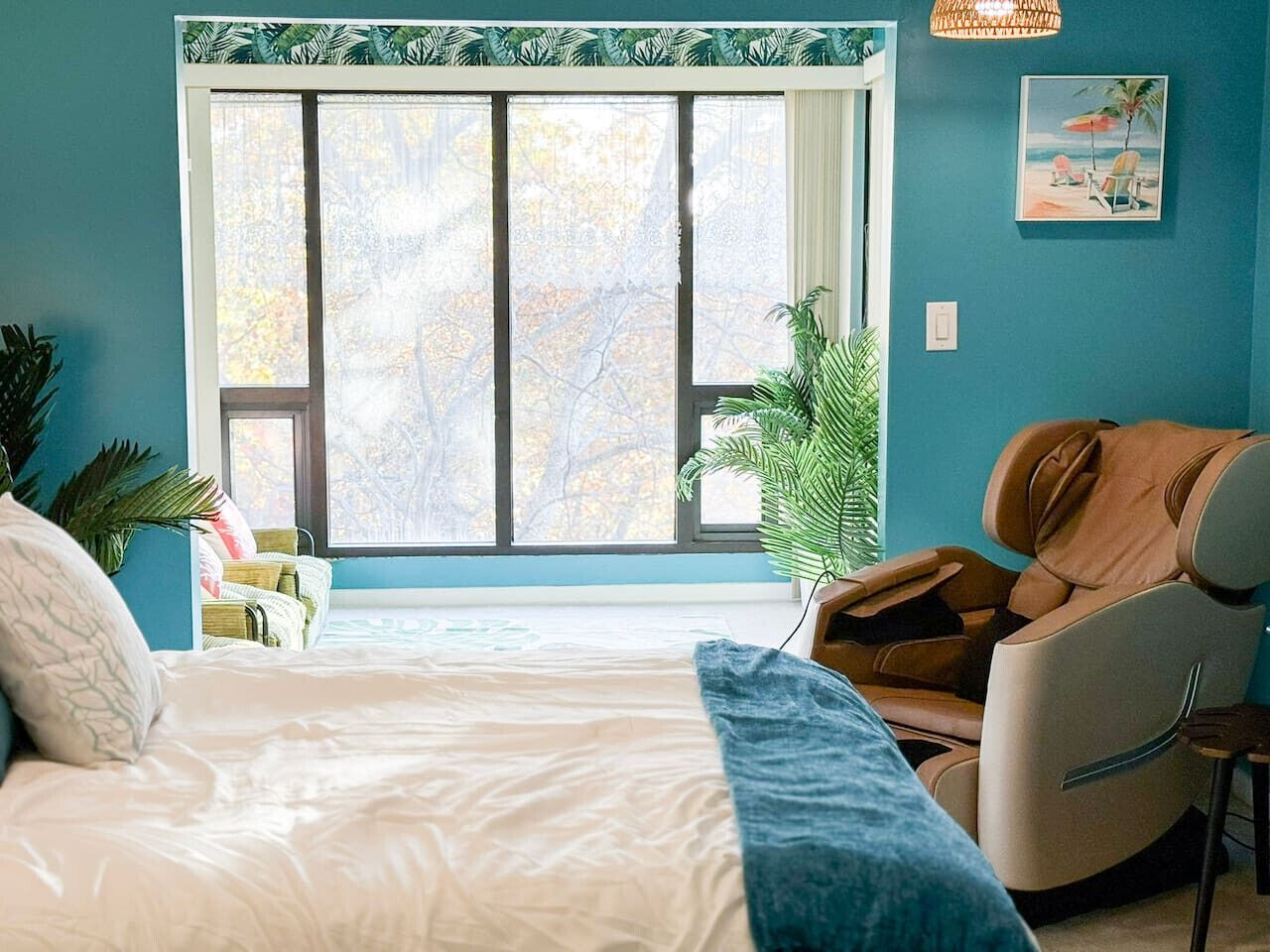 Un dormitorio con una cama grande, una silla de masaje, un ventanal con vistas a árboles y un cuadro con motivos tropicales en la pared.