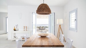 Un coin repas avec une table en bois, des chaises blanches, un luminaire suspendu et une vue sur l’extérieur grâce à une porte ouverte.
