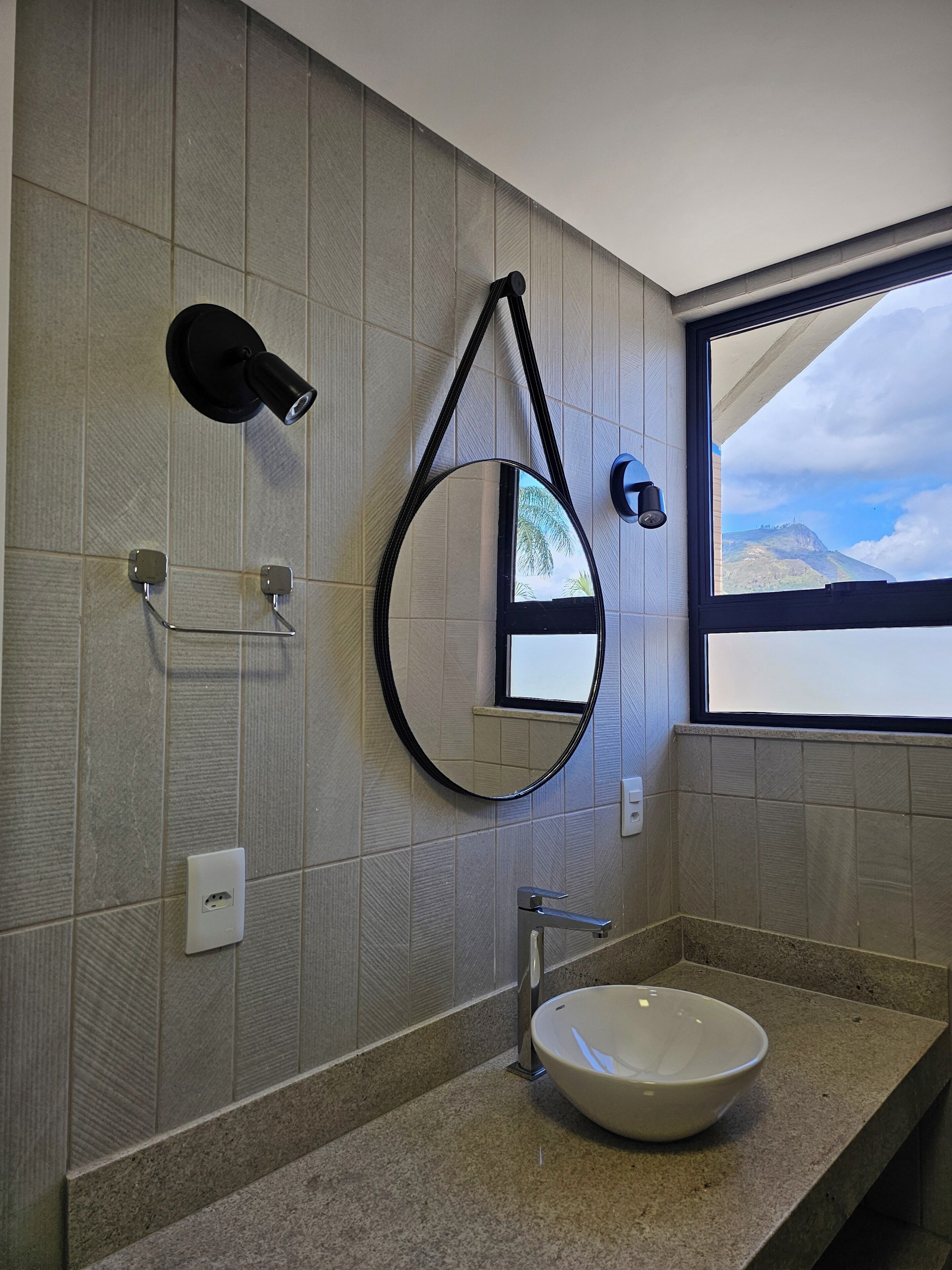 A bathroom with a round mirror, a wall-mounted light, a towel rack, a sink, and a window with a view of mountains.