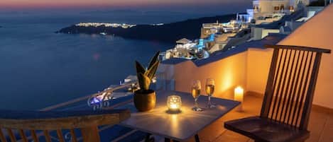 Terraço com mesa posta para dois, com vista para uma vila costeira ao anoitecer.
