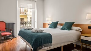 A hotel room with a bed, a chair, a nightstand, a lamp, and a view of the outside balcony. - Versailles | The Rentals Collection (San Sebastián/Donostia)
