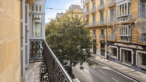 A narrow street with buildings on both sides, featuring balconies and a tree in the middle. - Versailles | The Rentals Collection (San Sebastián/Donostia)