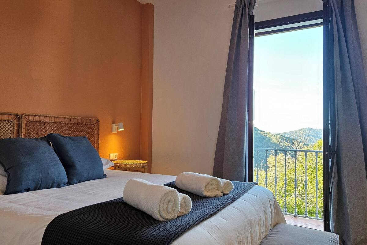 A hotel room with a bed, pillows, a nightstand, a lamp, and a view of the outdoors.