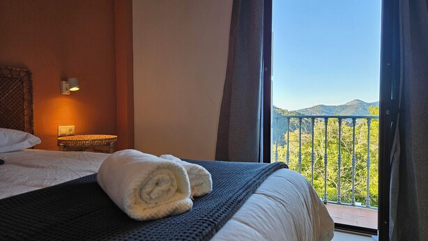 A hotel room with a bed, a bedside lamp, and a view of a balcony with a railing and greenery. - Hotel Rural Mirador de Jubrique (Jubrique)