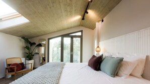 Een slaapkamer met een groot bed, houten plafond, een raam, een stoel en een plant.