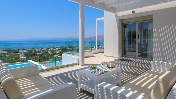 Een balkon met witte wicker meubels, een glazen tafel met een tijdschrift en een glas wijn, en uitzicht op zee en een verafgelegen landschap.