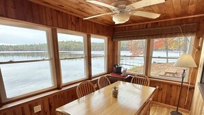 Una cabaña de madera con una mesa de comedor y sillas, ventanas amplias que dan a un lago y un ventilador de techo.