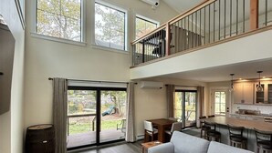 Een ruime woonkamer met een grote grijze modulaire bank, een eettafel en een keukenhoek. Er zijn meerdere ramen die zorgen voor natuurlijk licht en uitzicht op het buitenleven.