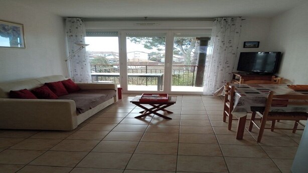 A living room with a sofa, a coffee table, and a dining table with chairs. There is a television on a stand and a balcony with a view of the outside.