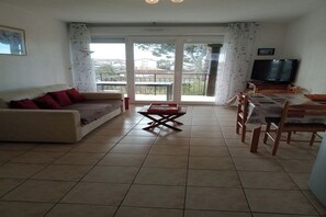 A living room with a sofa, a coffee table, and a dining table with chairs. There is a television on a stand and a balcony with a view of the outside. - Charming 2-room apartment in Soustons (Soustons)