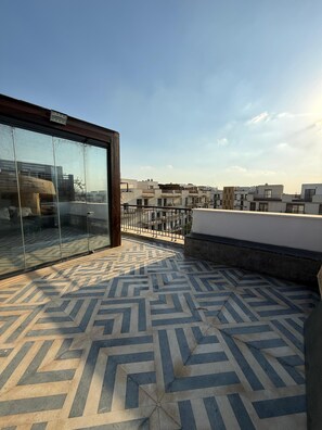 A rooftop terrace with a patterned floor, a glass wall, and a railing overlooking a residential area.