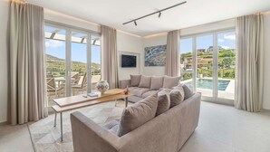 A modern living room with a beige sofa, a wooden coffee table, and a view of a pool and landscape through large windows. - Thrilling Andros Villa | Villa Merope | 2 Bedrooms | Private Outdoor Pool |. (Andros)