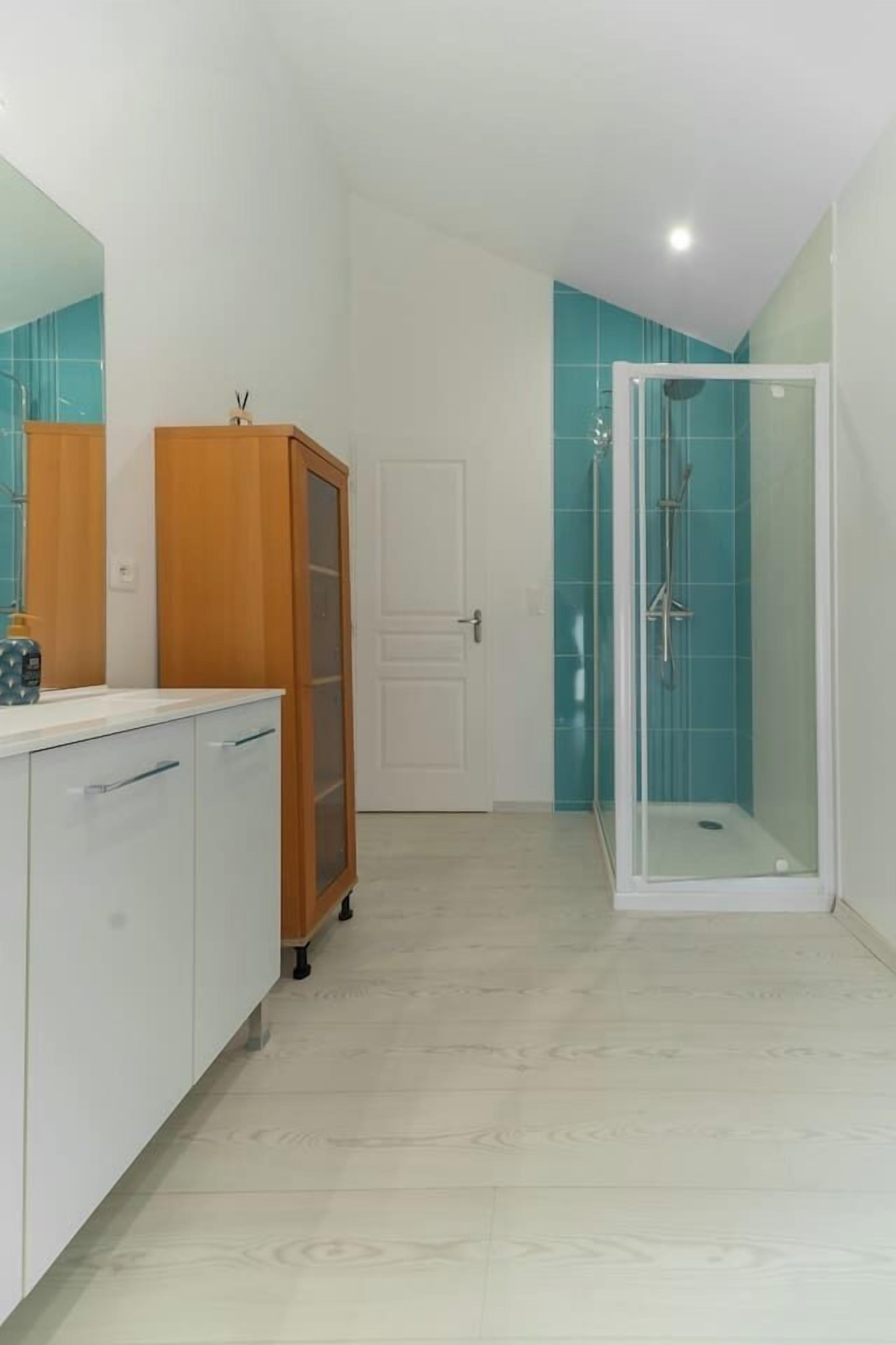 A bathroom with a white vanity, a wooden cabinet, a glass shower enclosure, and teal tiled walls.