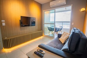 Ein modernes Wohnzimmer mit einer Couch, einem Couchtisch mit Fernbedienungen, einem an einer Holzwand montierten Fernseher und einer Glasschiebetür, die zu einem Außenbereich führt.