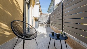 En smal udendørs terrasse med en sort stol, et lille rundt bord med to kopper og en potteplante.