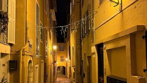 Uma rua estreita com edifícios de ambos os lados, com calçada de pedra, varandas e luzes de corda.