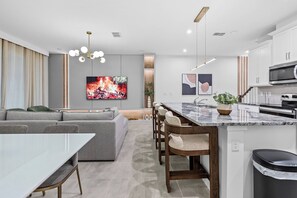 Eine moderne Küche mit Kochinsel, ein Essbereich mit Glastisch und ein Wohnzimmer mit Sofa und Fernseher.