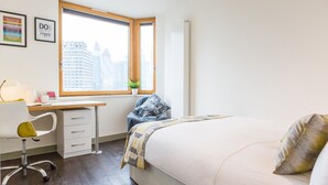 A bedroom with a bed, a desk, a chair, a window with a view of buildings, and a radiator. - Student Luxury Studio Aldgate Residence (London)