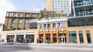 A multi-story building with a mix of modern and traditional architecture, featuring a ground-level commercial space with large windows and a variety of shops. - Student Luxury Studio Aldgate Residence (London)