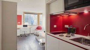 A modern kitchen with red walls, a stainless steel sink, and a window overlooking a cityscape. - Student Luxury Studio Aldgate Residence (London)