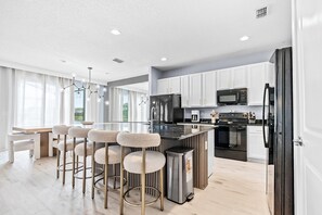 A modern kitchen with white cabinets, a stainless steel refrigerator, a microwave, and a coffee maker. There are four bar stools with a fluffy seat and a small trash bin. - 8BR house with private pool in Bella Vida Resort (Kissimmee)