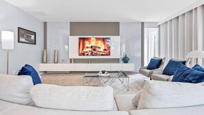 Ett modernt vardagsrum med en platt-TV som är monterad på en vit vägg, en beige soffa, ett soffbord i glas och en sidobord med dekorativa föremål.