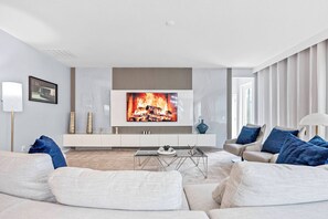 Ein modernes Wohnzimmer mit einem Flachbildfernseher an einer weißen Wand, einem beige gefärbten Sofa, einem Glastisch und einer Anrichte mit dekorativen Gegenständen.