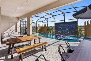 An outdoor pool area with a wooden table and benches, a sofa, and a large screen displaying a cityscape. - 10BR house sleeps 23 with game room (Clermont)