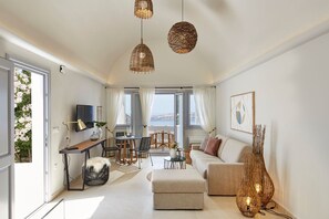 A modern living room with a sofa, ottoman, dining table, chairs, and a TV. There are wicker pendant lights, a plant, and a view of the outdoors. - Magnificent Santorini Villa | Villa Aphrodite’s Dream | 2 Bedrooms |. (Santorini)