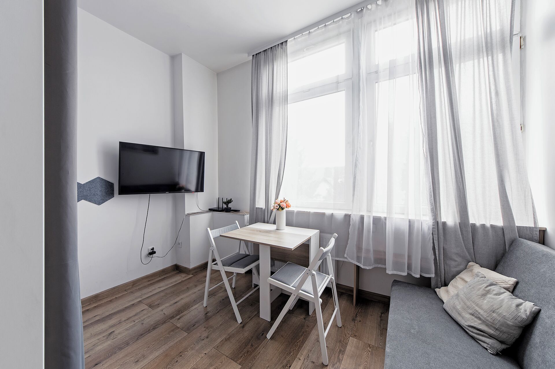Una camera moderna con TV a schermo piatto fissata alla parete, un tavolo in legno con due sedie, un divano grigio con un cuscino e tende leggere alla finestra.
