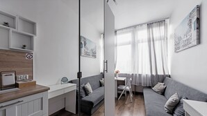 Een moderne, goed verlichte kamer met een grijze bank, een kleine eettafel en een witte kast. Er zijn planken boven de bank en een schilderij aan de muur.