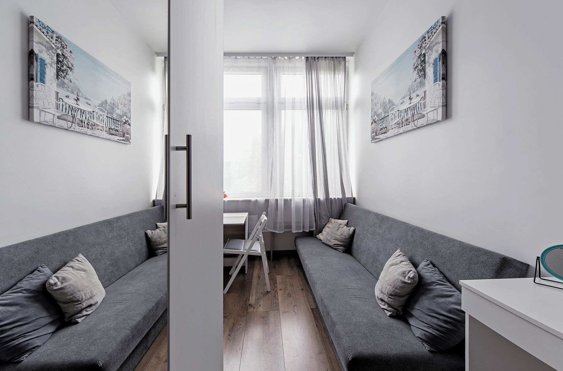 Een moderne woonkamer met twee grijze banken, een klein tafeltje en een wit bureau. Er hangen twee grote schilderijen aan de muren en er is een raam met dunne gordijnen.