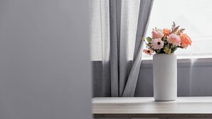 Een witte vaas met bloemen op een houten oppervlak, nabij een raam met grijze gordijnen.