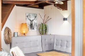 Ein modernes Wohnzimmer mit einem grauen Ecksofa, einem Holztisch, einer Wandlampe, einem Kaktus und einem Bild mit einem Reh.