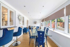 Ein Essbereich mit Glastisch und blauen Stühlen, große Fenster mit Blick auf einen Garten und eine durchgehende Tür erkennbare Küche.