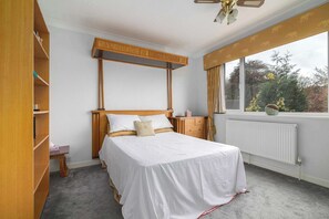 Ein Schlafzimmer mit Bett, Holz-Kopfteil, Nachttischen, Heizkörper und einem Fenster mit Blick auf Bäume.